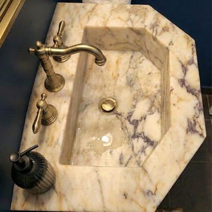 May include: A marble sink with gold taps and a soap dispenser. The sink has an octagonal shape and is made of beige marble with purple veining. A white towel hangs above the sink.