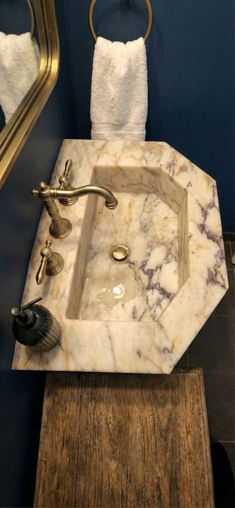 May include: A marble sink with gold taps and a soap dispenser. The sink has an octagonal shape and is made of beige marble with purple veining. A white towel hangs above the sink.