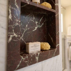 May include: A built-in marble shelf in a bathroom, featuring a deep red and white veined pattern. The shelf holds two bottles of shampoo, a bar of soap, and a natural sponge. The soap is labeled "ANYA ROSE GERANIUM".