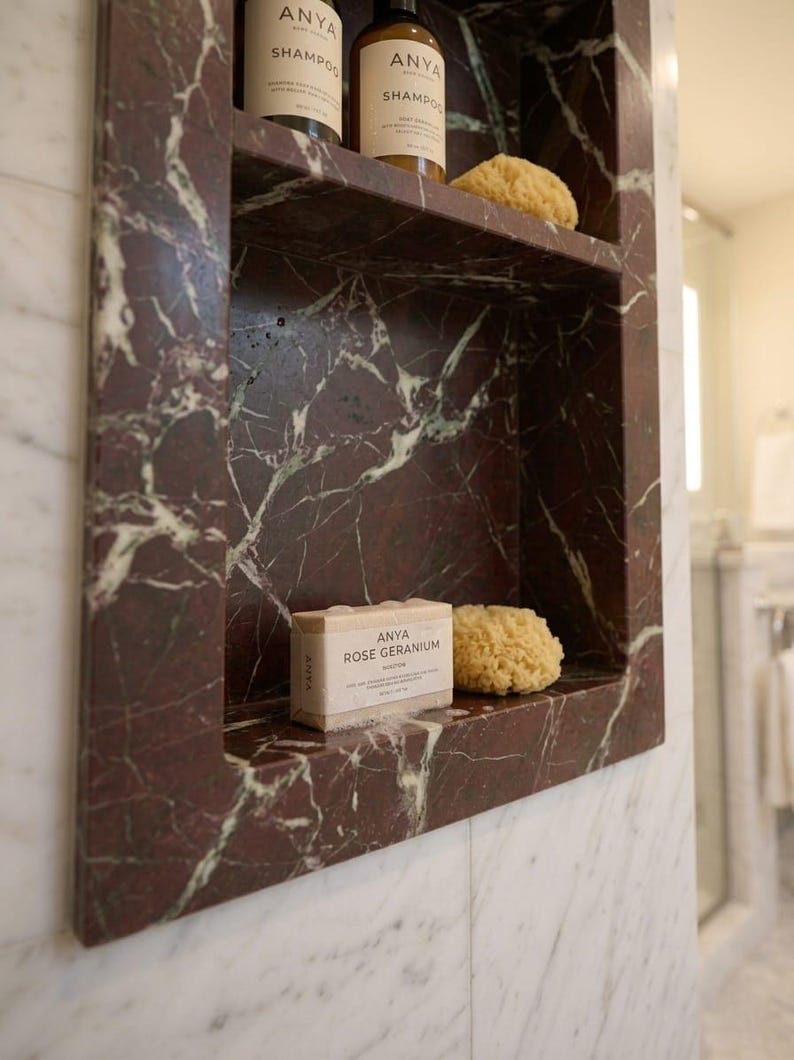 May include: A built-in marble shelf in a bathroom, featuring a deep red and white veined pattern. The shelf holds two bottles of shampoo, a bar of soap, and a natural sponge. The soap is labeled "ANYA ROSE GERANIUM".
