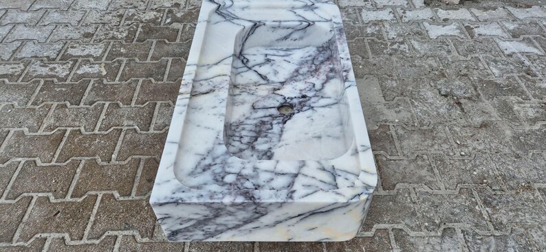 May include: A rectangular marble sink with a white base and intricate gray and purple veining. The sink has a rectangular basin and a smaller, recessed area. The sink is made of natural stone and is suitable for a bathroom.