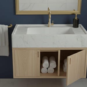 May include: A modern bathroom vanity with a white marble sink and a gold faucet. The vanity has light wood cabinets with rolled white towels inside. A gold towel bar holds a white towel. The background is a dark blue wall.