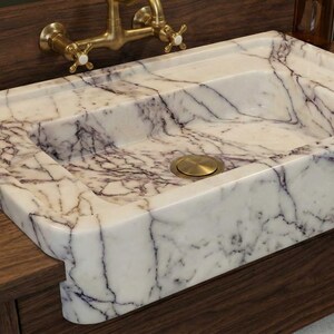 May include: Rectangular marble sink with a white base and purple veining. The sink has a brass drain and is set on a dark wood vanity. A brass faucet is mounted on the wall above the sink.