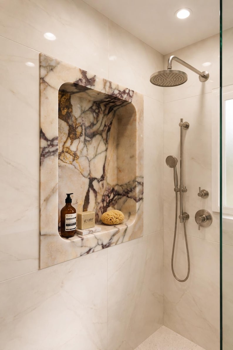 May include: A marble shower niche with a brown soap dispenser, a bar of soap, and a natural sponge. The niche is framed by a marble surround with purple veining. A shower head and handheld shower are visible.