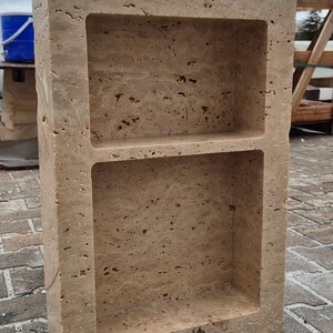 May include: A rectangular, beige stone shelf unit with two recessed compartments. The stone has a textured surface with small holes and variations in color. The unit is designed for storage or display.