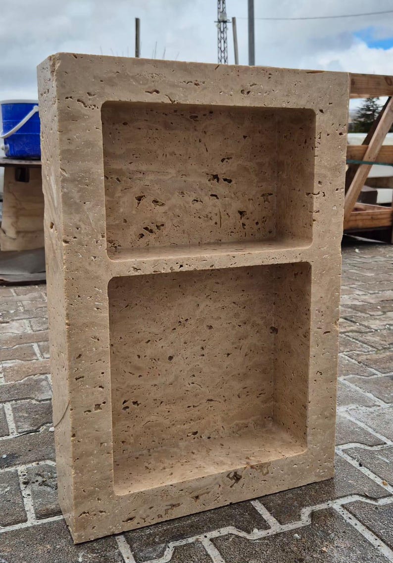 May include: A rectangular, beige stone shelf unit with two recessed compartments. The stone has a textured surface with small holes and variations in color. The unit is designed for storage or display.