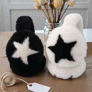 Op de afbeelding: Twee zachte oorbeschermers, één zwart met een witte ster en één wit met een zwarte ster. De oorbeschermers zijn gemaakt van een zacht, pluche materiaal. Een klosje touw en een blanco label staan op de voorgrond.