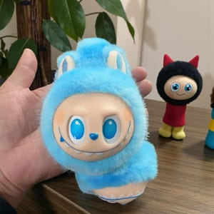 Pode incluir: Um brinquedo de pelúcia azul com um rosto sorridente e grandes olhos azuis. O brinquedo tem um capuz azul peludo com orelhas brancas e um corpo azul. Outras figuras são visíveis no fundo.