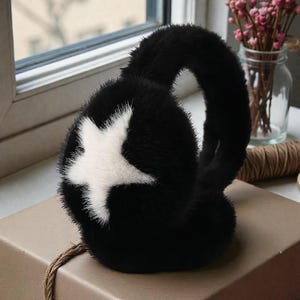 Op de afbeelding: Zwarte oorbeschermers met een wit sterontwerp. De oorbeschermers zijn gemaakt van een zacht, pluche materiaal. De ster is gecentreerd op de oorbeschermer. De oorbeschermer is verbonden door een zwarte band.