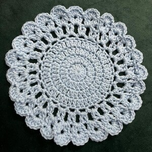 May include: A round, light blue crochet doily featuring a dense, spiraling center pattern that transitions into an openwork design with a scalloped, shell-like border. The piece is crafted from yarn and displays a consistent, textured stitch throughout.