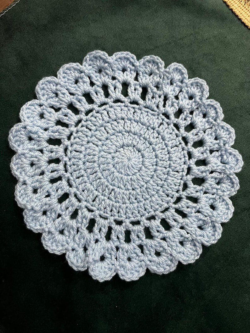 May include: A round, light blue crochet doily featuring a dense, spiraling center pattern that transitions into an openwork design with a scalloped, shell-like border. The piece is crafted from yarn and displays a consistent, textured stitch throughout.