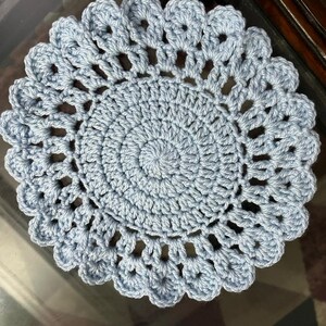 May include: A round, light blue crochet doily featuring a dense, circular center pattern that transitions into an open, airy stitch design. The outer edge is finished with a scalloped border, creating a decorative, circular textile piece.