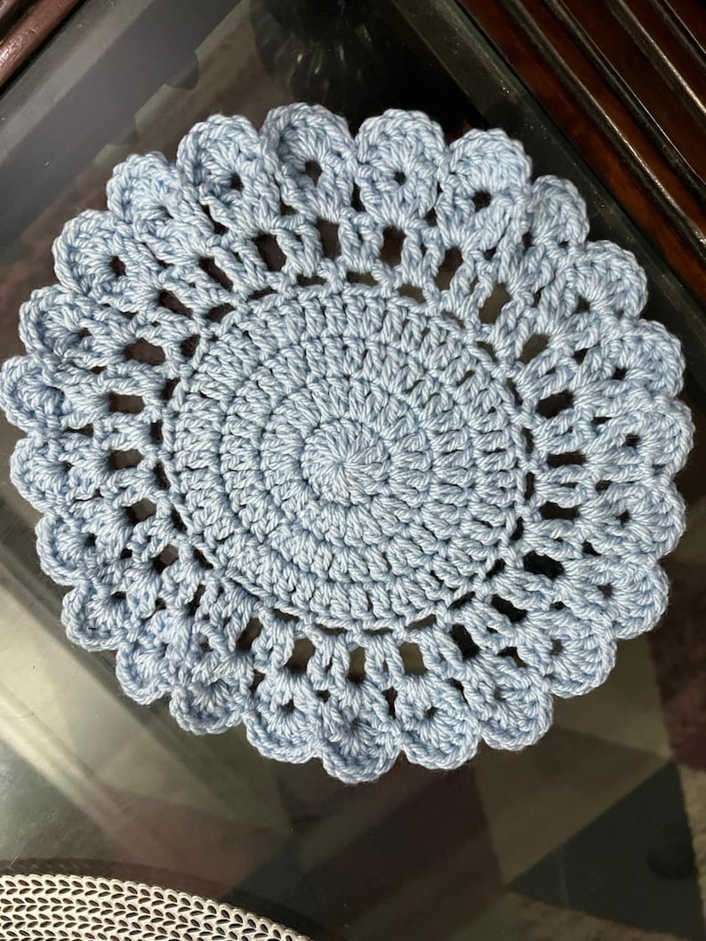 May include: A round, light blue crochet doily featuring a dense, circular center pattern that transitions into an open, airy stitch design. The outer edge is finished with a scalloped border, creating a decorative, circular textile piece.