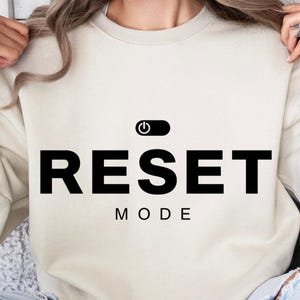 May include: A cream-colored sweatshirt with the words "RESET MODE" in bold black letters. Above the word "RESET" is a black power button symbol. The sweatshirt is a casual style.