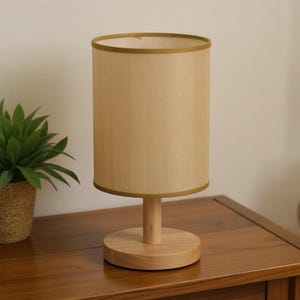 Peut inclure: Une lampe de table beige avec un abat-jour cylindrique et une base en bois. L'abat-jour a un aspect texturé et est bordé d'une bande dorée. La lampe est posée sur une table en bois, avec une plante en pot visible.