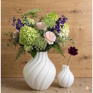 K&ouml;nnte beinhalten: Zwei wei&szlig;e Keramikvasen mit einem strukturierten, wirbelnden Design. Eine Vase enth&auml;lt einen Strau&szlig; aus rosa Rosen, gr&uuml;nen Hortensien und lila Blumen. Eine einzelne dunkelviolette Blume befindet sich in der kleineren Vase. Der Hintergrund ist eine hellbraune Holzoberfl&auml;che.