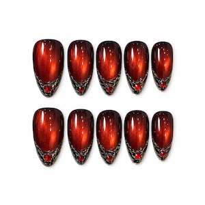 May include: A set of ten press-on nails featuring a deep, metallic red cat-eye finish. Each nail has a pointed almond shape and is decorated at the base with a textured, dark metallic silver filigree pattern and a single small, round red rhinestone.