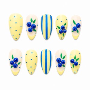 May include: A set of ten almond-shaped press-on nails featuring a yellow and blue color palette. Designs include yellow nails with blue polka dots, yellow nails with blue vertical stripes, and cream-colored nails adorned with 3D blue berry accents.