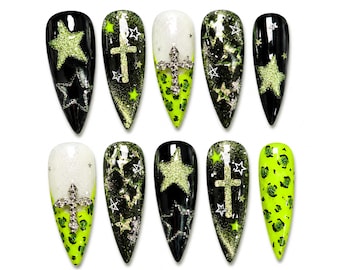 Op de afbeelding: Een set van tien stiletto-vormige kunstnagels met zwarte, witte en neongroene ontwerpen. Patronen bevatten glittersterren, metalen kruisbedels en een neongroene luipaardprint. De nagels hebben een glanzende afwerking met diverse stermotieven.