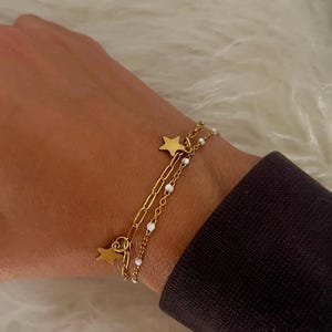 Pode incluir: Conjunto de pulseiras douradas com duas correntes. Uma corrente tem pingentes de estrela e a outra pequenas contas brancas. As pulseiras são usadas em um pulso, em um fundo branco e macio.
