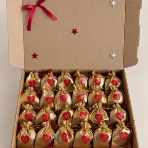 May include: A cardboard box containing twenty-four small gold drawstring bags, each featuring a red heart with a number from 1 to 24. The inside lid is decorated with a red ribbon bow, one red star, and two silver stars.