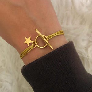 Peut inclure: Bracelet doré avec un charm en forme d'étoile et un fermoir à bascule. Le bracelet est composé de plusieurs brins de cordon doré. Le charm en forme d'étoile est suspendu à un anneau relié au fermoir.