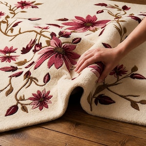 May include: A cream-coloured area rug featuring a tufted floral pattern in shades of deep pink, magenta, and brown. The design consists of winding vines, leaves, and blooming flowers spread across the surface, showcasing a thick, plush texture.