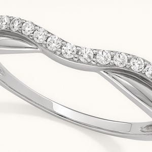 May include: A silver-colored ring with a curved band, set with a row of small, clear gemstones. The ring's design features a split band that crosses over itself, creating an elegant and modern look. The gemstones sparkle.
