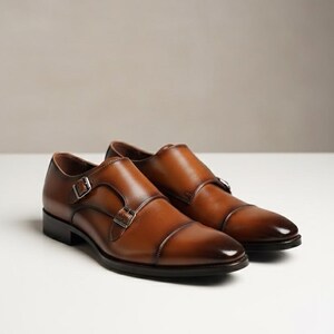 May include: A pair of brown leather double monk strap shoes. The shoes feature a polished finish, silver buckles, and a classic cap-toe design. The shoes are displayed on a white surface.