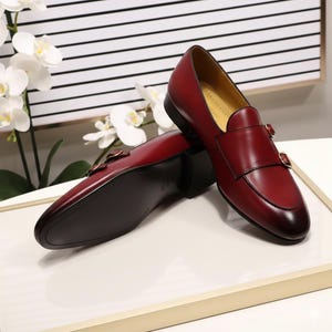 Pu&ograve; includere: Un paio di scarpe eleganti in pelle bordeaux con doppia fibbia. Le scarpe hanno un design elegante con suola scura. Sono esposte su una superficie bianca. Misurano circa 28 cm di lunghezza.