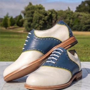 May include: A pair of two-tone oxford shoes featuring cream-coloured pebbled leather and smooth blue leather overlays. The shoes include white laces, yellow contrast stitching along the brogue detailing, and a light brown stacked wooden heel and sole.