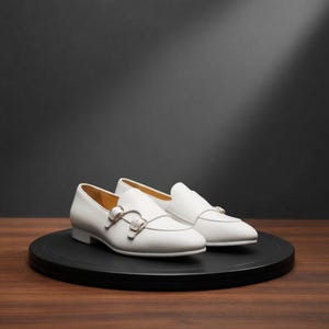 May include: A pair of white leather dress shoes with double monk straps and gold-colored buckles. The shoes are displayed on a black circular platform against a dark background. The shoes have a low heel and a pointed toe.