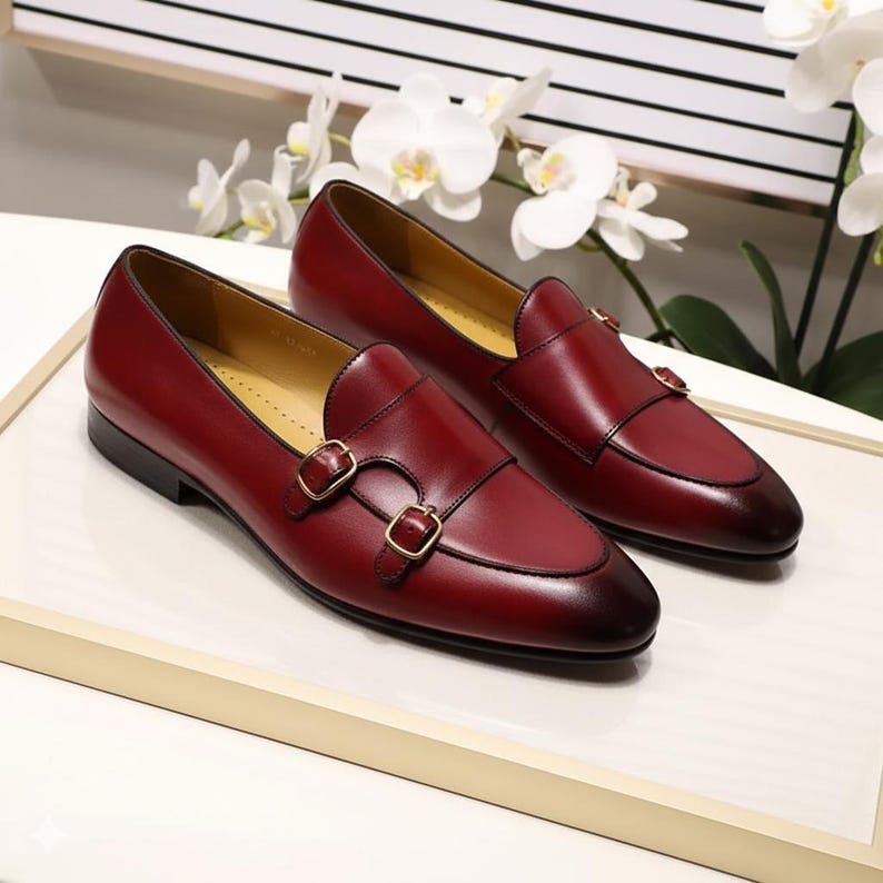 Pu&ograve; includere: Un paio di mocassini in pelle bordeaux con chiusure a doppia fibbia. Le scarpe hanno una finitura lucida e un puntale marrone scuro. L'interno &egrave; di una tonalit&agrave; pi&ugrave; chiara di marrone. Le scarpe sono esposte su una superficie bianca.