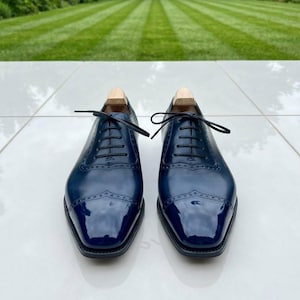 May include: A pair of polished navy blue leather oxford shoes featuring a cap toe with decorative brogue detailing, black laces, and light-coloured wooden shoe trees inserted into the heels, set against a clean, reflective white surface.