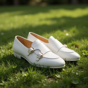 May include: A pair of white leather shoes with gold-colored double buckles, set on a bed of green grass. The shoes have a classic design with a pointed toe and a low heel, suitable for formal or casual wear.