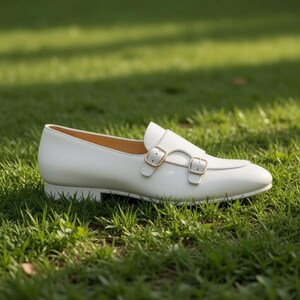 May include: A white leather shoe with a double monk strap design. The shoe features two gold-colored buckles and a low heel. The shoe is set against a backdrop of green grass.