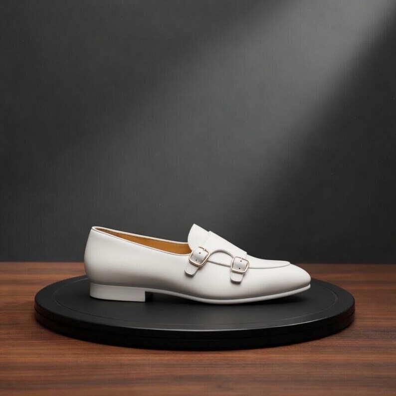 May include: A white leather loafer with a double monk strap buckle closure. The shoe is displayed on a black circular platform, set against a dark background. The shoe has a low heel and a sleek, minimalist design.