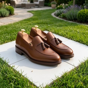 May include: A pair of brown leather loafers featuring a tassel detail on the vamp and a stitched apron toe. Each shoe includes a light-colored wooden shoe tree with a gold-toned metal knob inserted into the interior to maintain the shape.