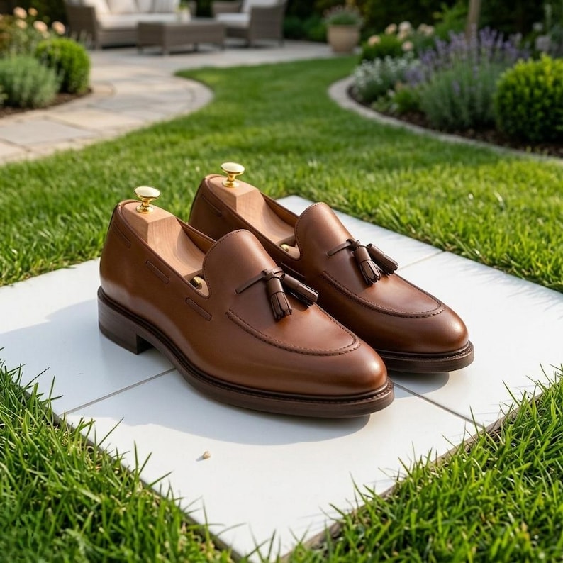May include: A pair of brown leather loafers featuring a tassel detail on the vamp and a stitched apron toe. Each shoe includes a light-colored wooden shoe tree with a gold-toned metal knob inserted into the interior to maintain the shape.
