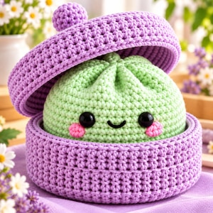 May include: A light green crocheted dumpling character with black eyes and pink cheeks, nestled inside a round, purple crocheted container with a matching lid that has a small pom-pom on top, crafted with visible yarn stitches.