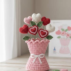 May include: A handmade crochet bouquet of heart-shaped flowers in a pink vase tied with a white ribbon. The bouquet includes red, pink, and white hearts with green leaves. A card with a similar design is in the background.