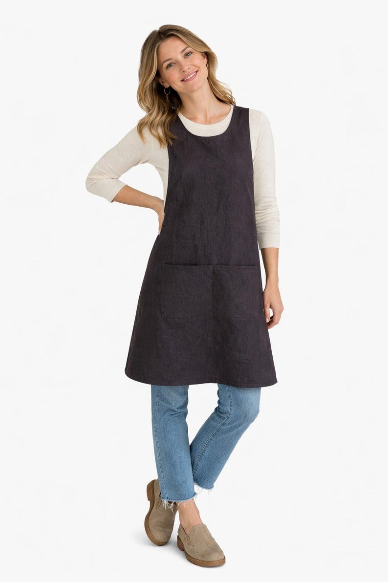 K&ouml;nnte beinhalten: Eine dunkelgraue Sch&uuml;rze mit zwei gro&szlig;en Vordertaschen. Die Sch&uuml;rze wird &uuml;ber einem cremefarbenen Langarmshirt und einer blauen Jeans getragen. Die Sch&uuml;rze ist aus einem strukturierten Stoff gefertigt.