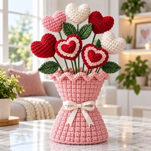 May include: A crochet bouquet of heart-shaped flowers in red, pink, and white, featuring green leaves on stems. The flowers are arranged in a textured, light pink crocheted vase with a ruffled top edge and a decorative white ribbon tied in a bow.