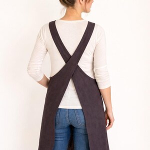 K&ouml;nnte beinhalten: Eine dunkelbraune Sch&uuml;rze mit Kreuz-R&uuml;cken-Design. Die Sch&uuml;rze wird &uuml;ber einem wei&szlig;en Langarmshirt und einer blauen Jeans getragen. Die Tr&auml;ger der Sch&uuml;rze kreuzen sich am R&uuml;cken.
