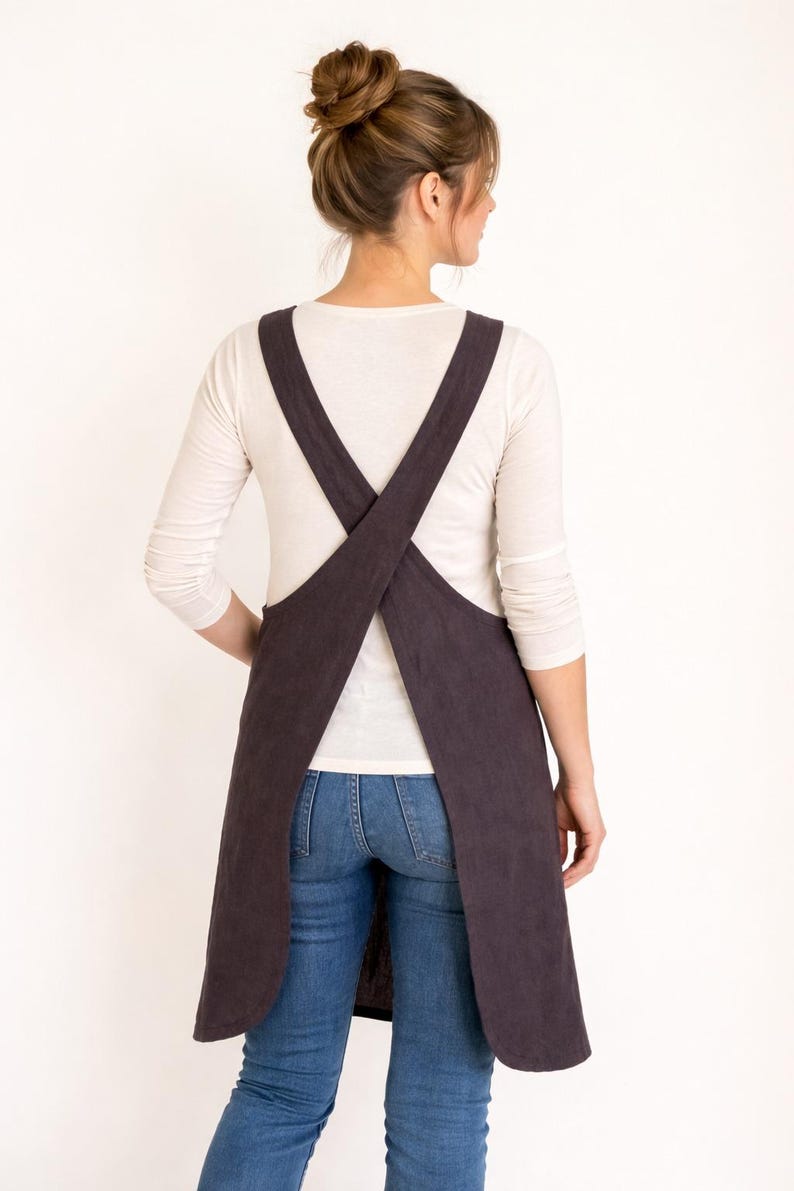 K&ouml;nnte beinhalten: Eine dunkelbraune Sch&uuml;rze mit Kreuz-R&uuml;cken-Design. Die Sch&uuml;rze wird &uuml;ber einem wei&szlig;en Langarmshirt und einer blauen Jeans getragen. Die Tr&auml;ger der Sch&uuml;rze kreuzen sich am R&uuml;cken.