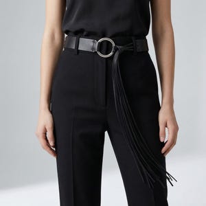 Peut inclure: Une ceinture en cuir noir avec une grande boucle circulaire argentée et de longues franges pendantes. La ceinture est portée avec un pantalon noir et un haut noir, créant un look monochrome. Le design de la ceinture ajoute un accent élégant.