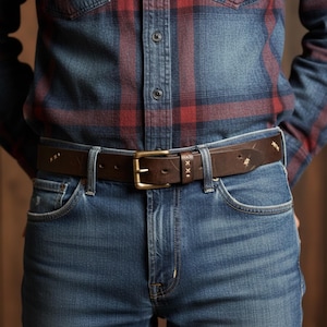 Peut inclure: Une ceinture en cuir marron avec une boucle en laiton est portée avec un jean bleu. La ceinture a des coutures décoratives et est fixée avec une boucle rectangulaire en laiton. Le jean est en denim délavé moyen, et la personne porte une chemise à carreaux.