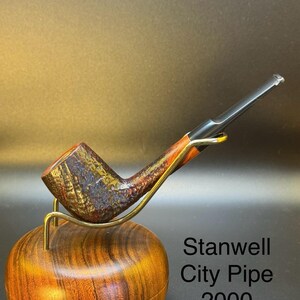 May include: A Stanwell City Pipe 2000 tobacco pipe with a dark, textured bowl and a black stem. The pipe rests on a wooden stand with a gold-colored metal support. The text "Stanwell City Pipe 2000 Hagebro Kro" is visible.