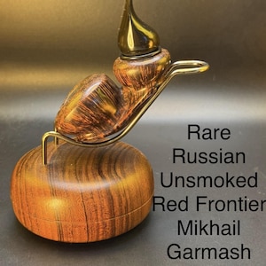 May include: A rare, unsmoked Red Frontier Mikhail Garmash Reverse Calabash pipe. The pipe is made of wood with a dark brown and reddish-brown grain pattern. It rests on a gold-colored metal stand and a wooden base. The text on the image reads: Rare Russian Unsmoked Red Frontier Mikhail Garmash Reverse Calabash.
