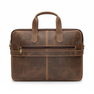 May include: A brown leather briefcase with a top handle and a front zipper pocket. The bag has a rectangular shape and a distressed finish, with visible stitching and a brass-colored zipper. The bag is designed for carrying documents and a laptop.